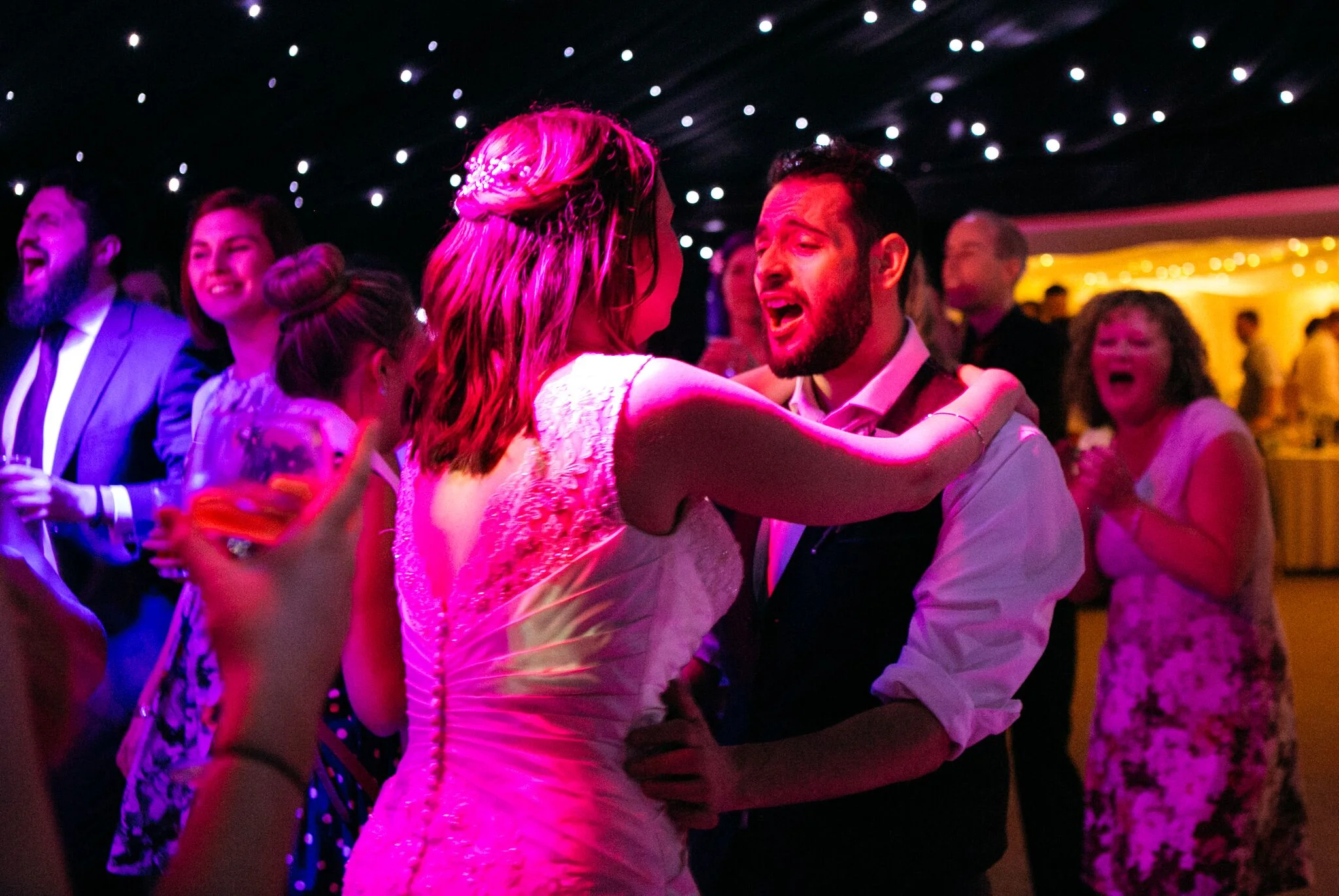 The bride and groom dancing together among their guests beneath pink party lighting