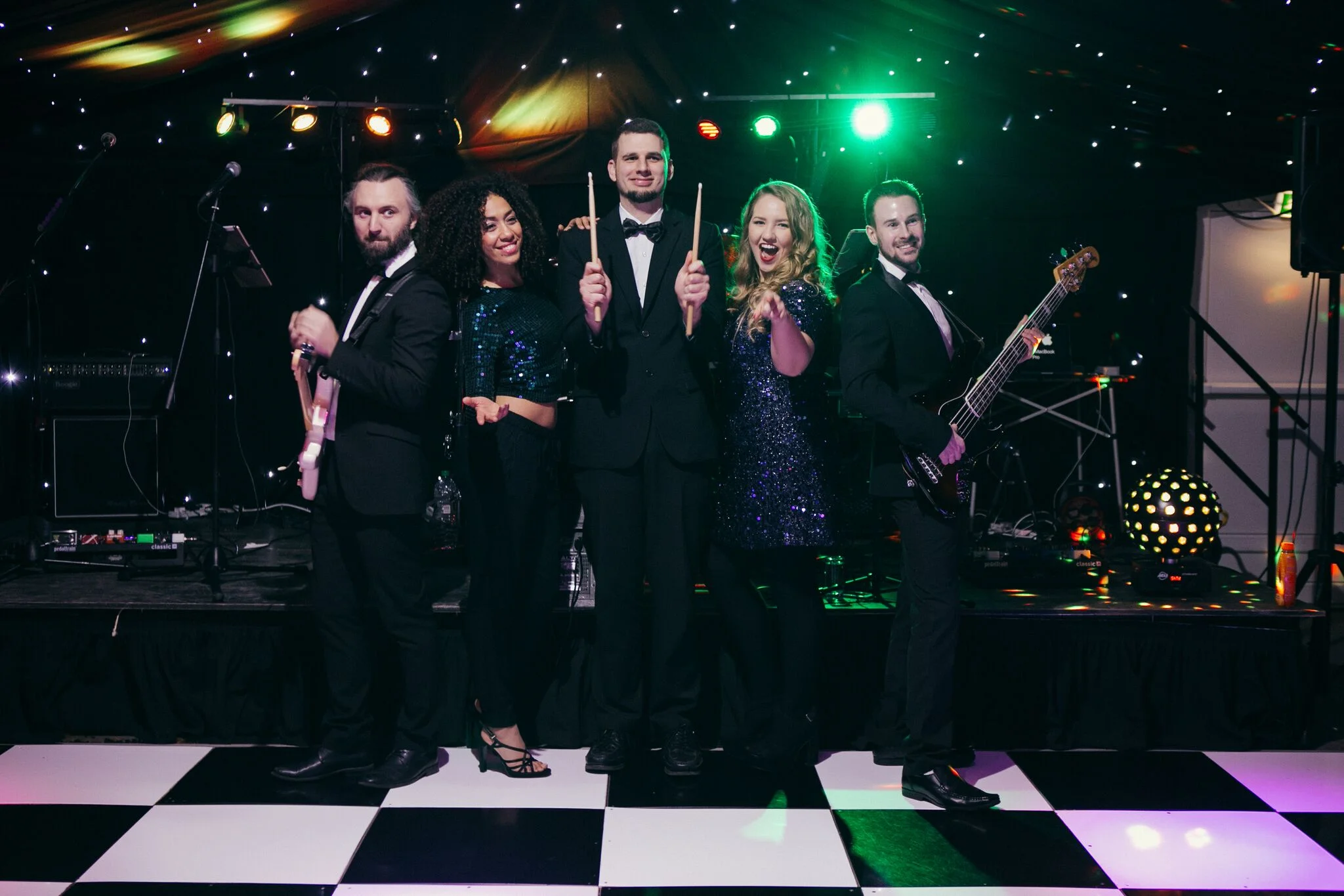 Two vocalists, a guitarist, a bassist and a drummer posing onstage beneath coloured lights and a star-lit backdrop
