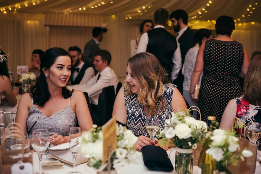 Wedding guests chatting at tables with white flowers beneath the marquee’s warm fairy lights