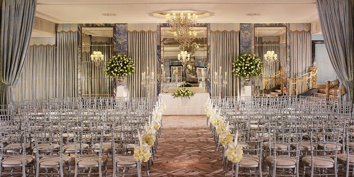 Wedding ceremony setup at The Dorchester with a white-flower-lined aisle, silver chairs and blue-and-gold drapes