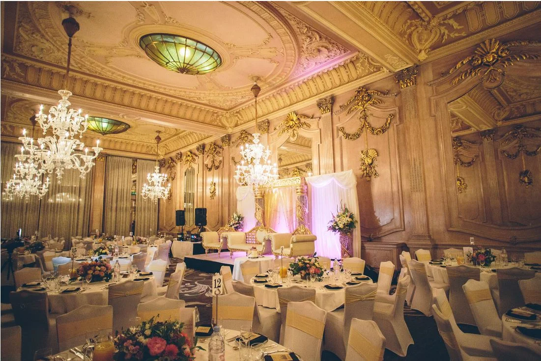 Ornate gold-toned reception room with chandeliers, round dining tables and a decorated wedding stage at the former Le Méridien Piccadilly
