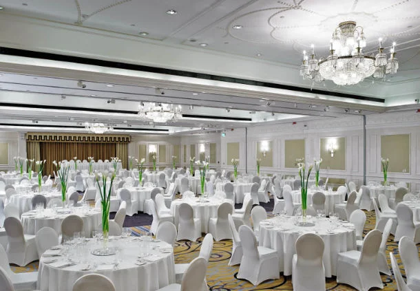 Round wedding breakfast tables with white chair covers and tall green flower arrangements in the Marriott hotel’s chandelier-lit room