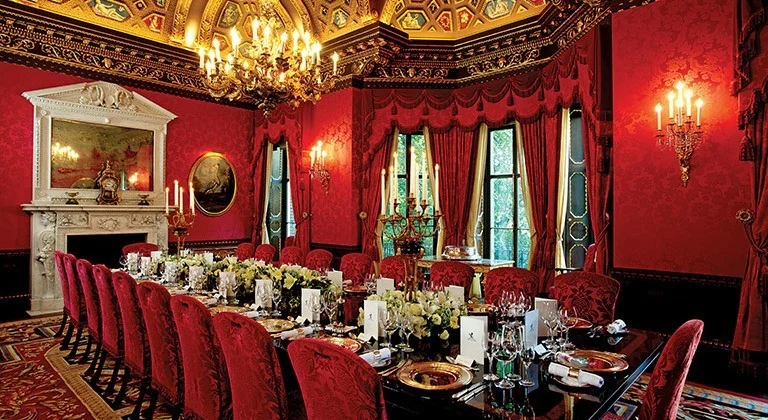 Long formal dining table surrounded by red upholstered chairs in an ornate red-and-gold private room at The Ritz