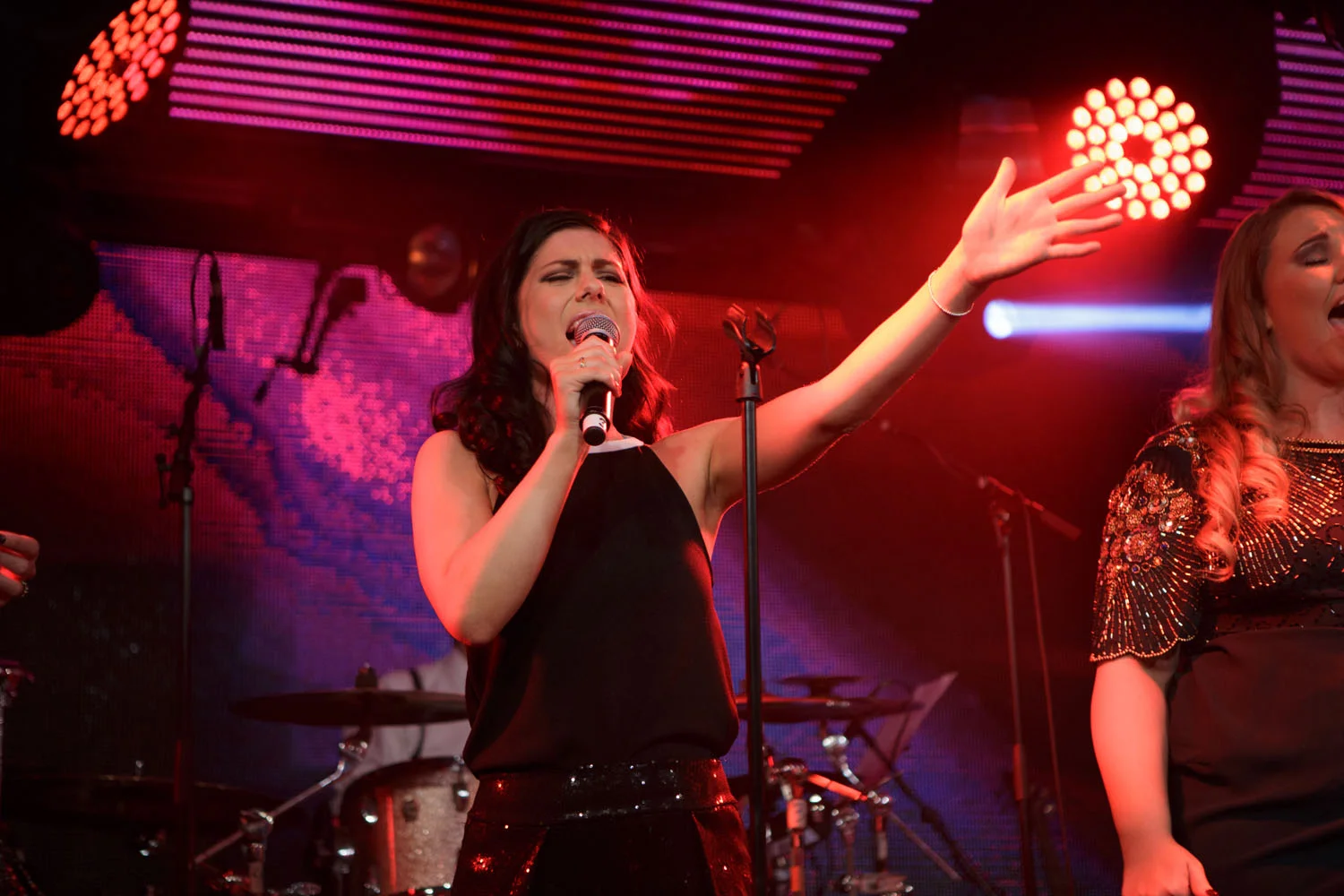 Phoebe singing into a handheld microphone with one arm raised, alongside another vocalist under red stage lighting